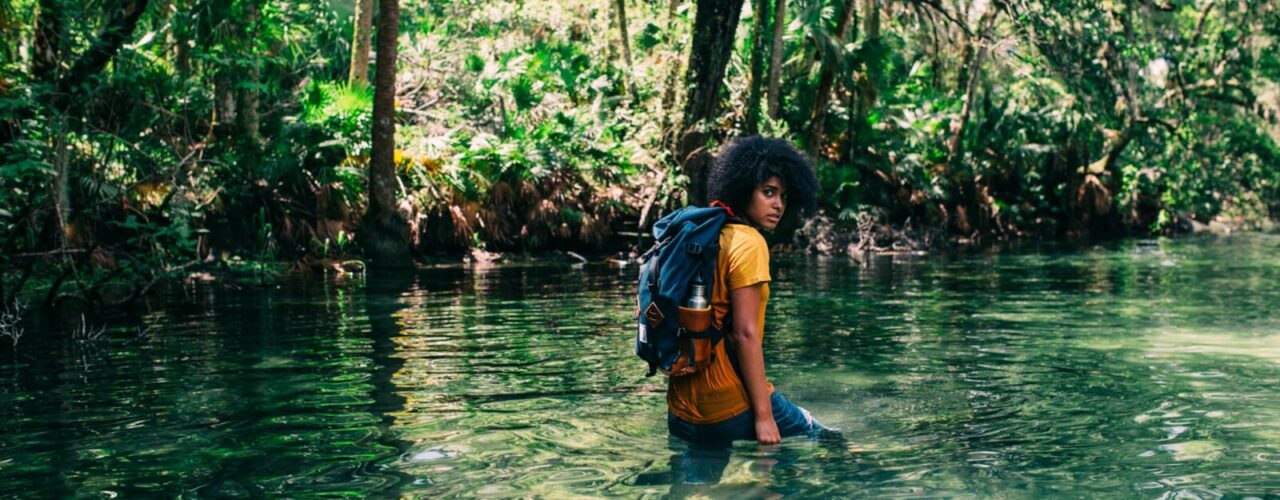 person in orange top wearing backpack walking on body of water in forest during daytime