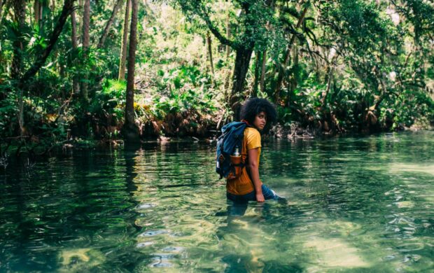 person in orange top wearing backpack walking on body of water in forest during daytime